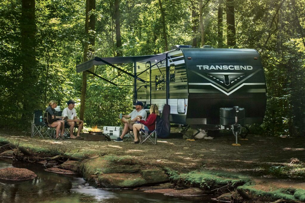 A Winnebago Transcend Travel Trailer parked in a wooded campsite and a family sitting around a fire pit outside.
