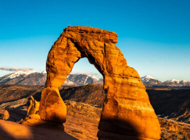 A red rock arch glows in the sun with mountains in the background with Utah landscapes as a great place to camp.