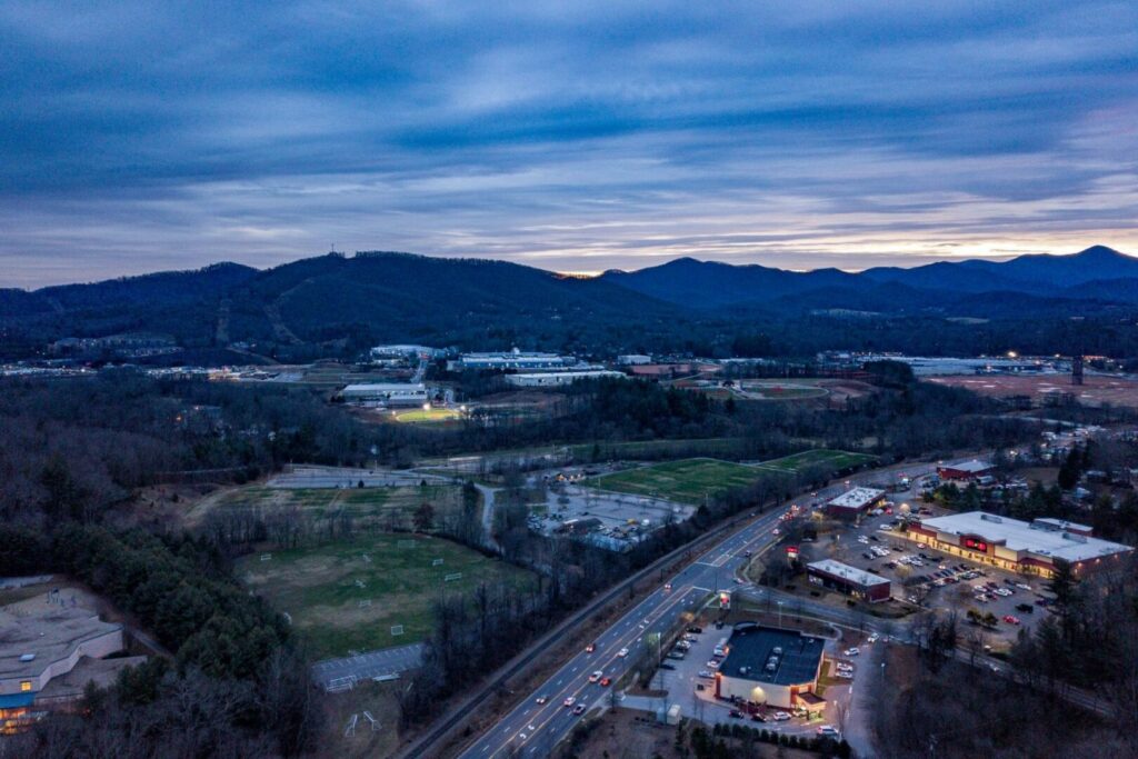 An aerial view of Asheville, NC, at dusk with the city lights starting to shine and the Blue Ridge Mountains in the distance.