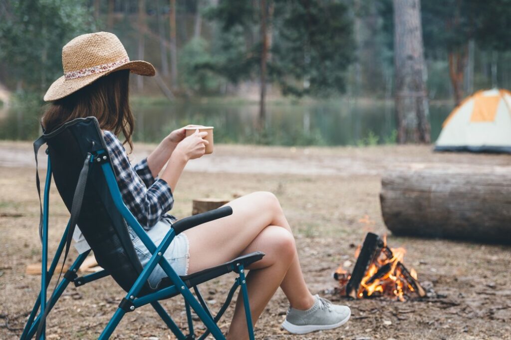 A woman relaxes in a camp chair next to a fire drinking out of a mug.