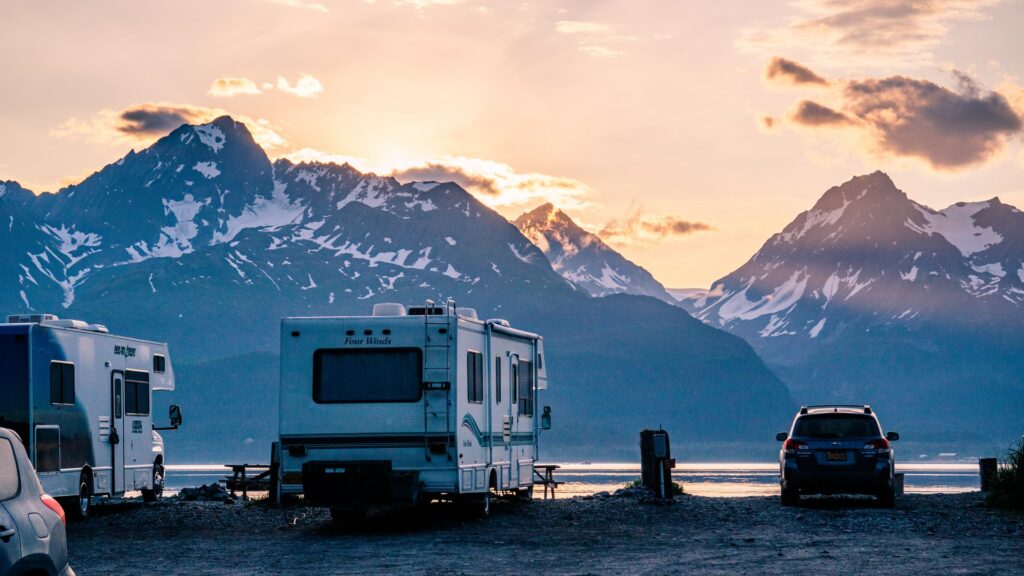 Rvs parked next to a lake looking out at tall, snowcapped mountains with the sun rising over them.