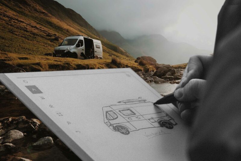 Drawing a conceptual van on a notepad with a class b van camping in the distance near a stream