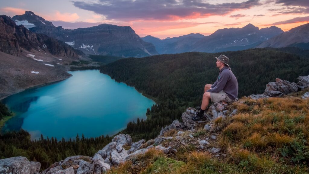 A man overlooks a beautiful glass lake in the Rocky Mountains as the sun colors the sky pink.