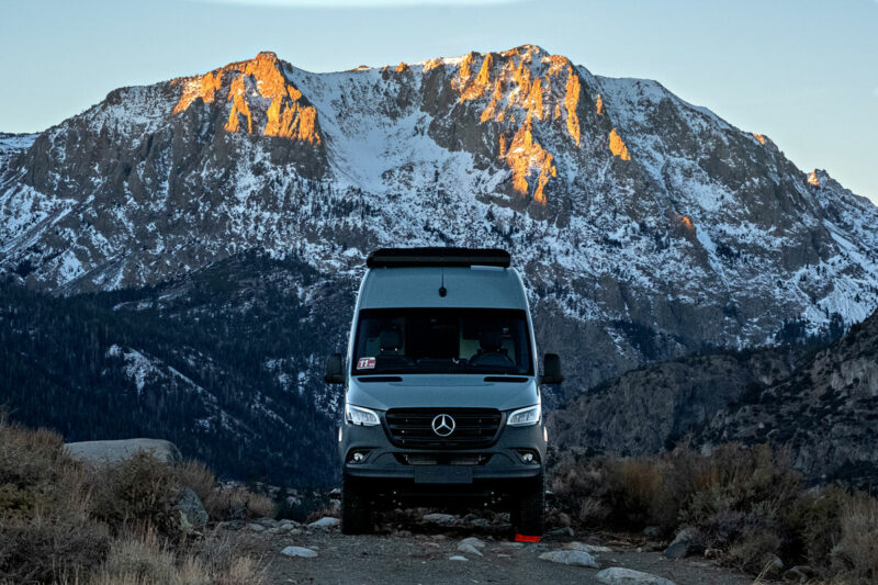 A Sprinter camper van parked in the mountains with snowcapped peaks behind it.