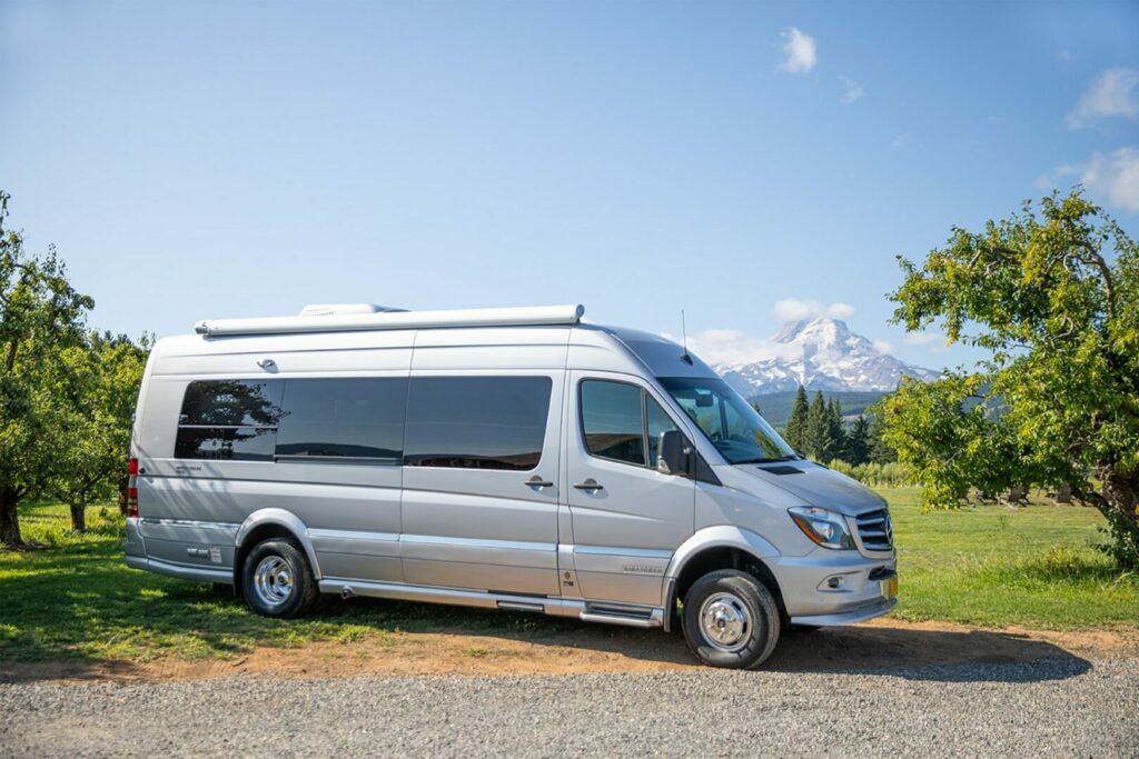 A new gray Mercedes Sprinter van parked along a gravel road with a mountain scenery behind it.