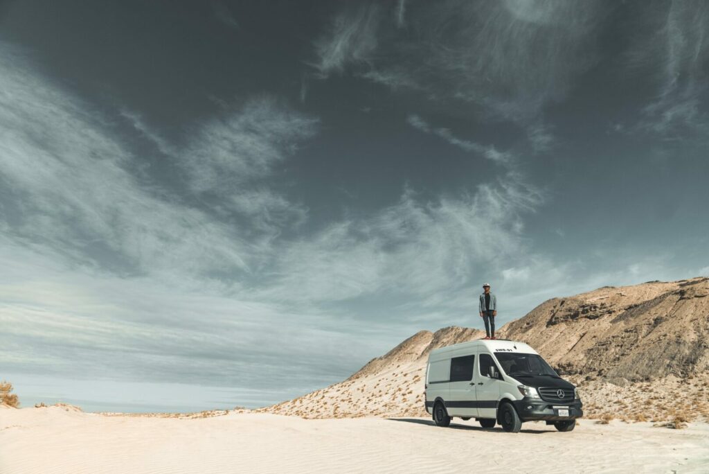 A van lifer stands on top of his camper van out in the desert.
