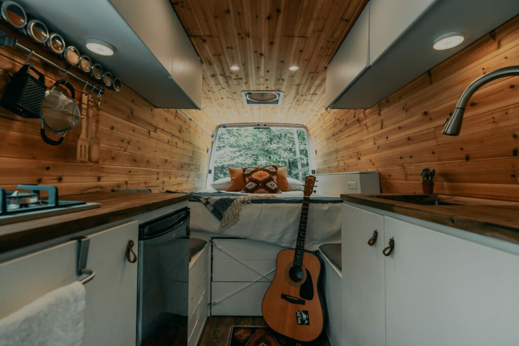 Inside view of a camper van with white cabinets, pine walls and ceiling, and a bunk in the back.