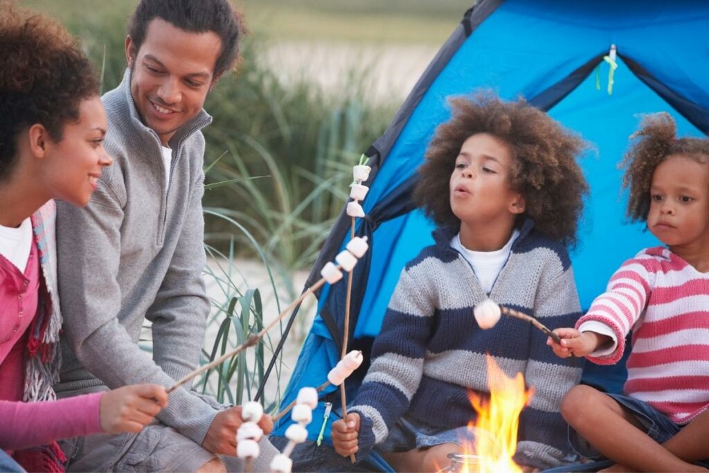 A family enjoys roasting marshmallows over a fire beside their camping tent.