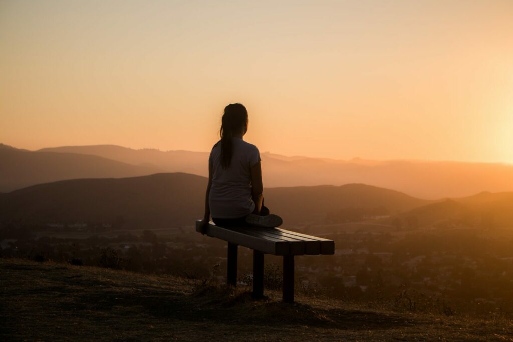 The silhouette of a woman resting on a bench, overlooking a sunset in the hills.