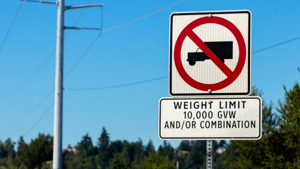 A sign on the side of a road that shows a truck with a red line through it and the words WEIGHT LIMIT. You need to know your GVWR when you see signs like these. 