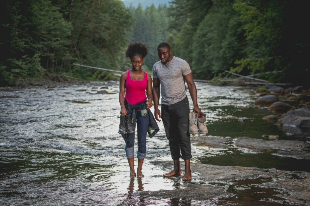 A couple walks barefoot in a river running through a pine forest.