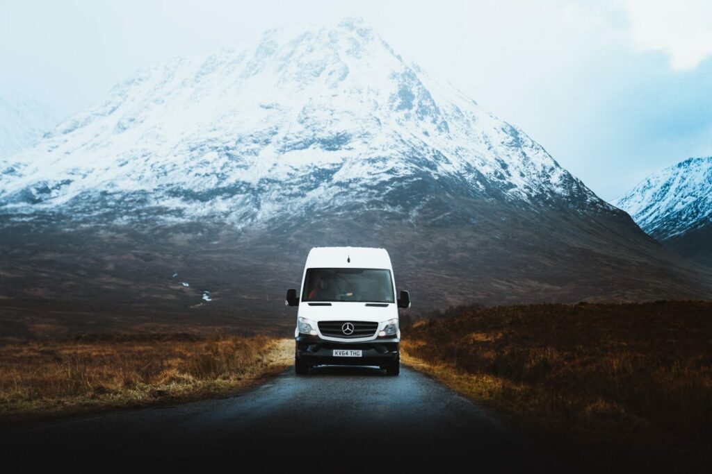 A Class B RV drives down a remote road with a snowcapped mountain behind it during the winter where a one night freeze is imminent.