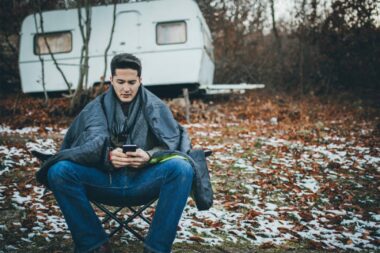 A man uses his phone outside while camping on a cold morning where snow has coated the ground around his travel trailer.