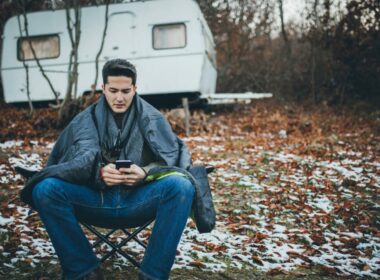 A man uses his phone outside while camping on a cold morning where snow has coated the ground around his travel trailer.