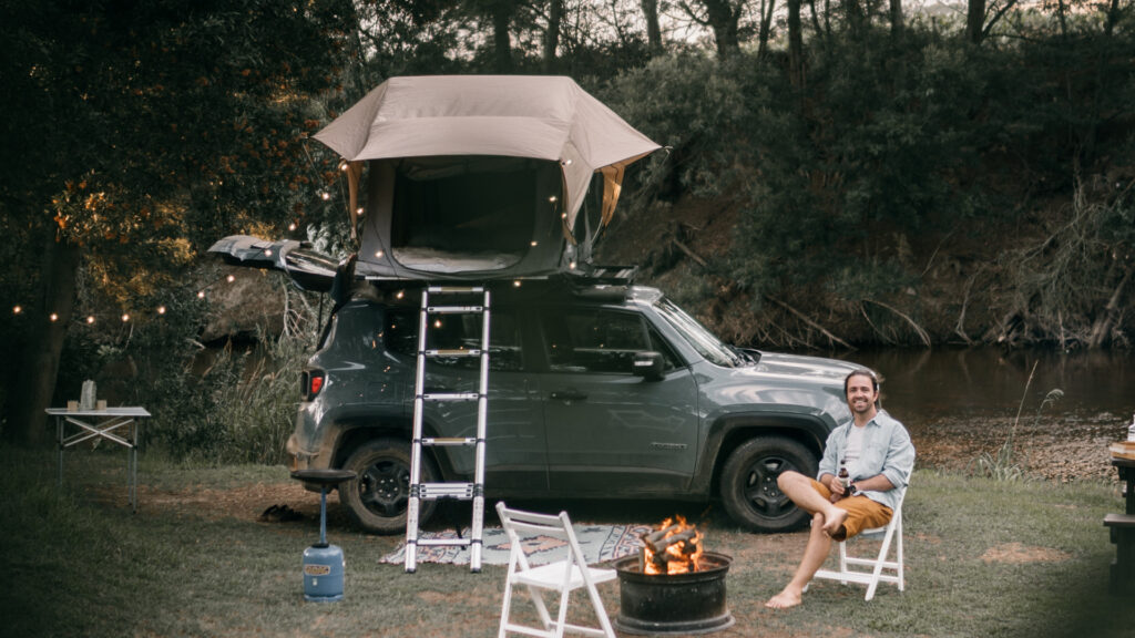 A smiling man sitting outside his jeep camper setup on top of his roof next to a river with a campfire going