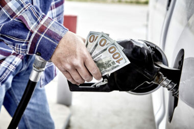Man's hand holding three hundred US dollars and gas nozzle while pumping gas into parked vehicle