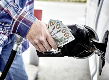 Man's hand holding three hundred US dollars and gas nozzle while pumping gas into parked vehicle