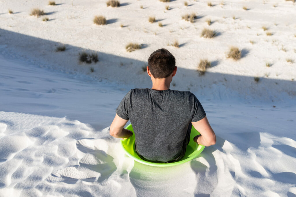 Back of man sitting on sand in white sands dunes national monument in New Mexico on green disk sled for sliding down hills