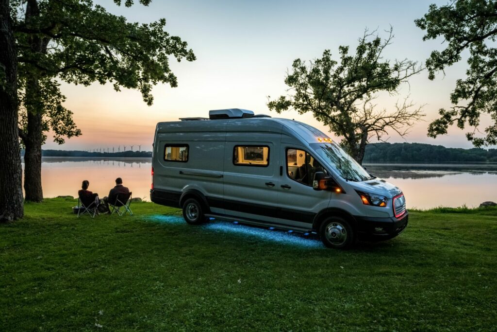 A couple sitting outside by a lake with their electric RV parked next to them 