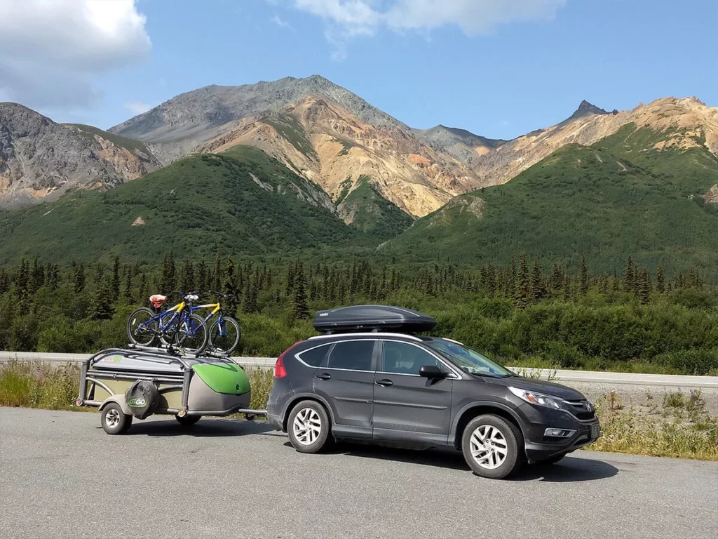 A sedan car pulling a micro camper with two bikes pulled over on the side of the road with mountains in the background