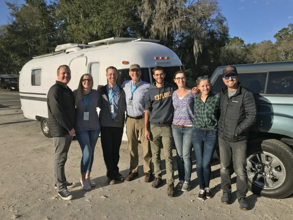 A group of RVing friends standing on front of a travel trailer at a rally