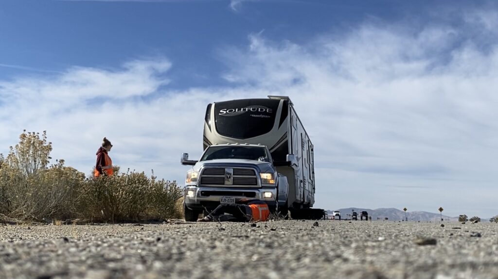 A man and woman in orange vests changing a tire on the side of the road while their fifth wheel is connected to their truck