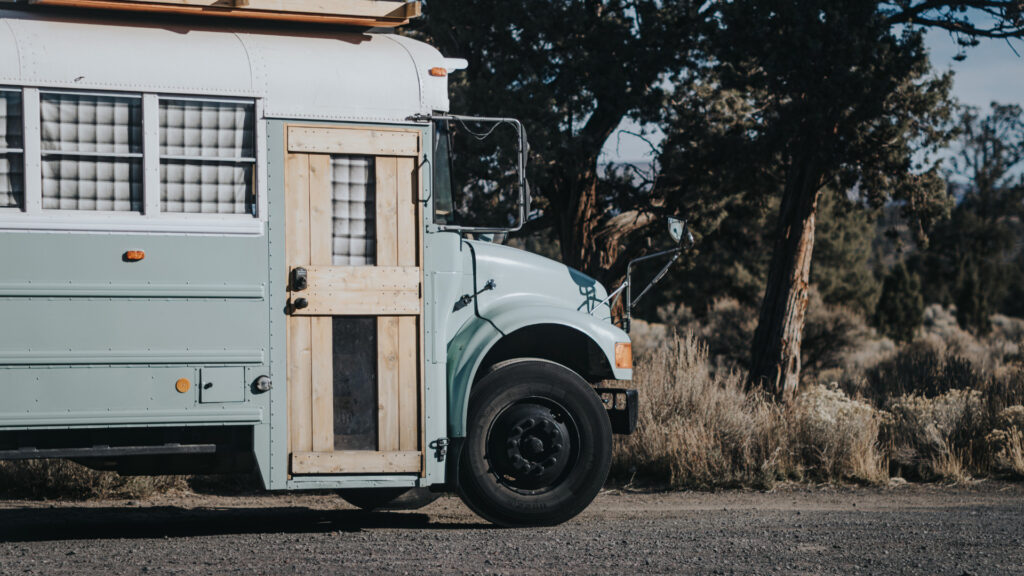 The front of a blue skoolie bus with a custom door parked on a dirt road.