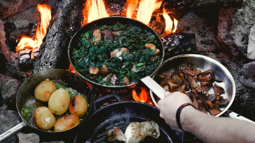 A man using his campfire cooking kit over an open flame to make potatoes, mushrooms, spinach, and meat. 