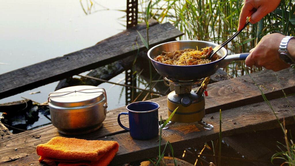 A person cooking stir fry in their campfire cooking kit next to a lake with grass in the background 