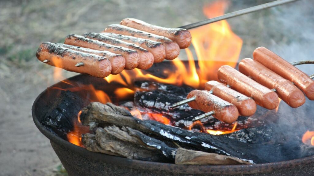 Two skewers loaded with hotdogs over a campfire. One of the camping hacks to try in 2022 is using a muffin tin for all your condiments. 