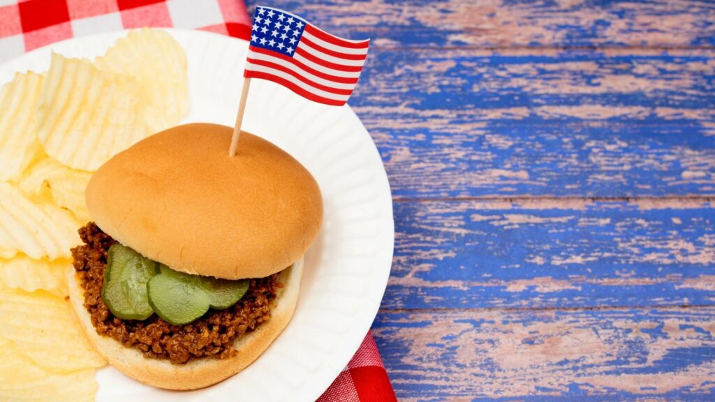 A sloppy joe sitting on a paper plate with an American flag toothpick through it.