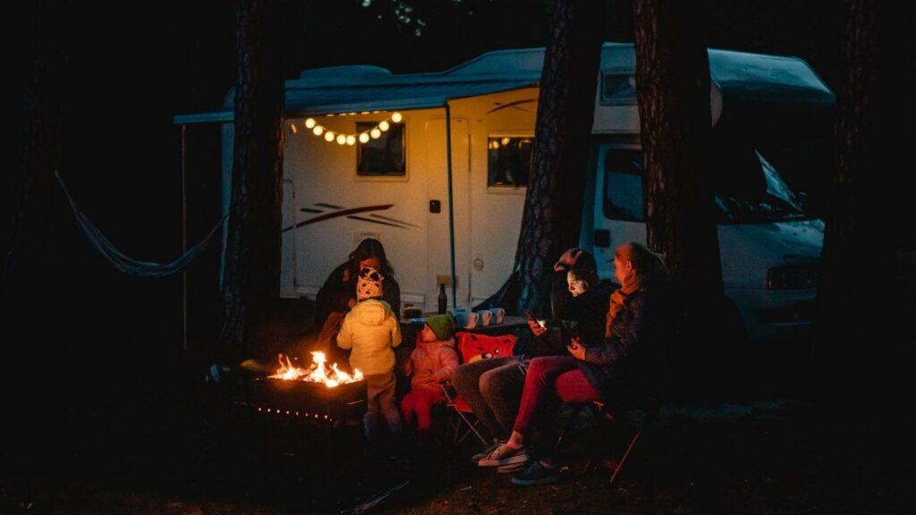 A family at night around a campfire with RV string lights hanging from their camper awning in the background 