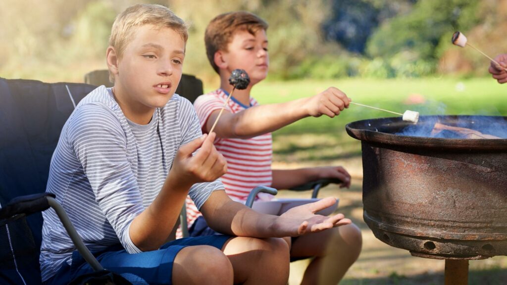 A boy looking at his burnt marshmallow on a stick that he cooked in the campfire next to him 