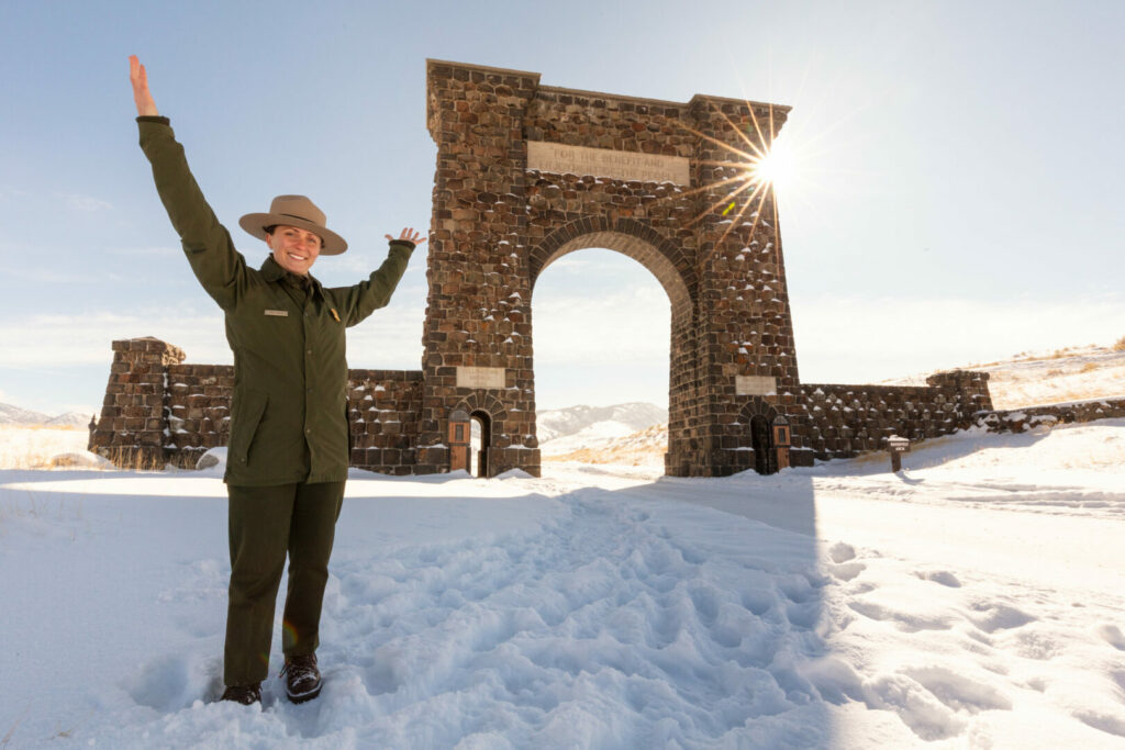 Volunteer ranger with her arms up at the opening gate of Yellowstone which is covered in snow.
