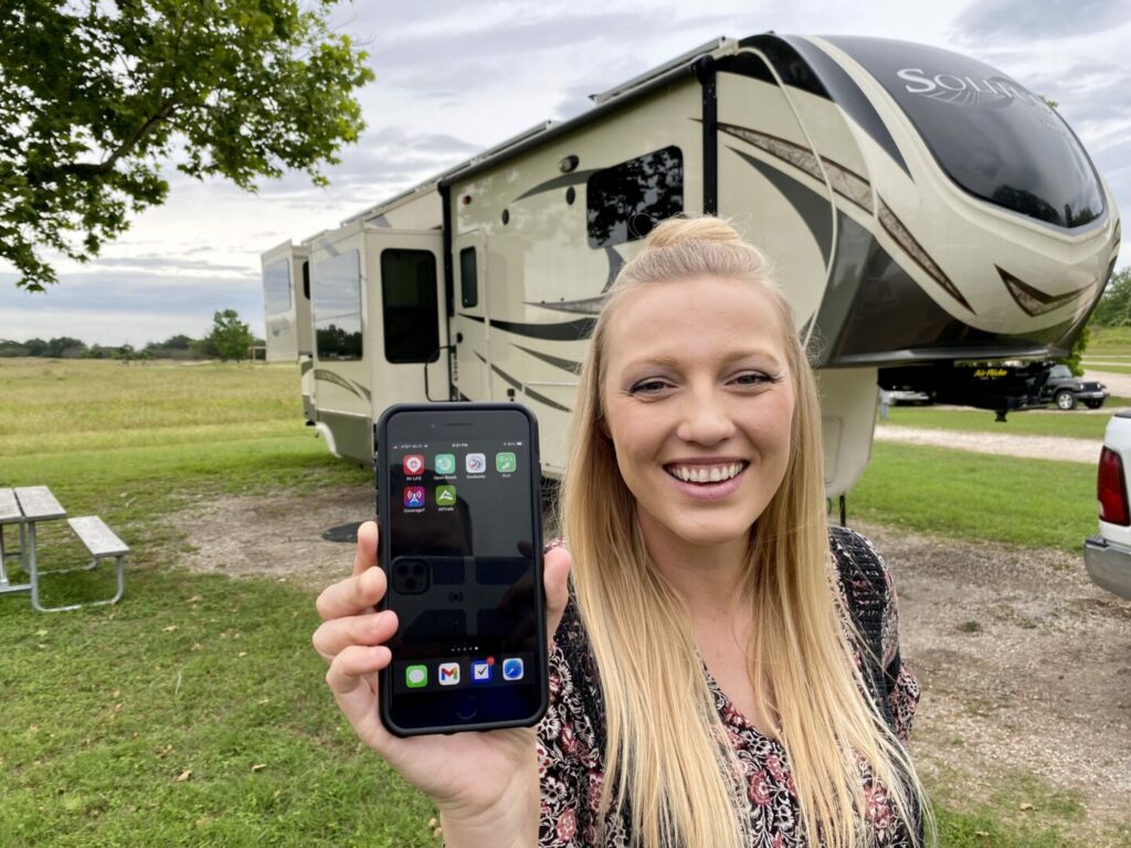 A woman holding a phone showing the latest apps for RVing in front of her fifth wheel RV