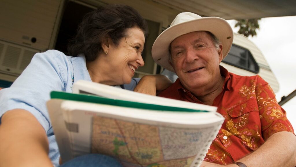An older couple sitting in front of an RV smiling at each other and holding a map 