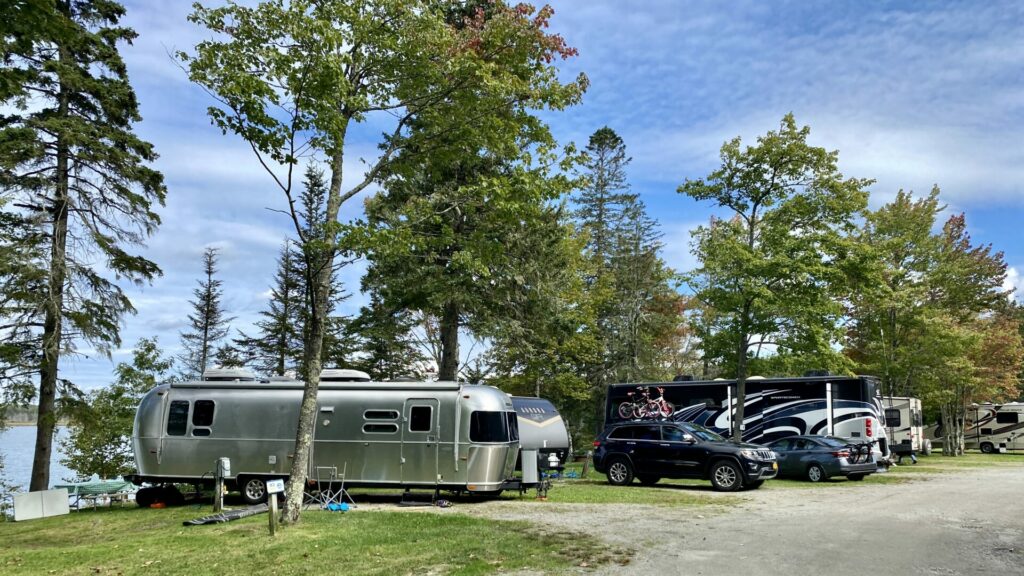A row of RVs in a campground in Maine with a river right behind the sites and trees 