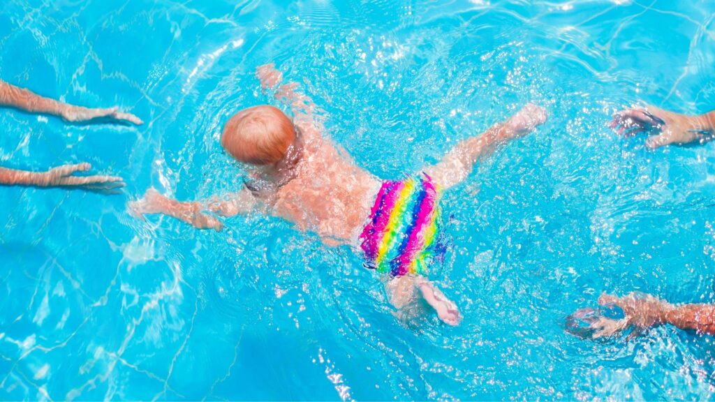 A baby swimming in a pool from one set of hands to another with a swim diaper on.