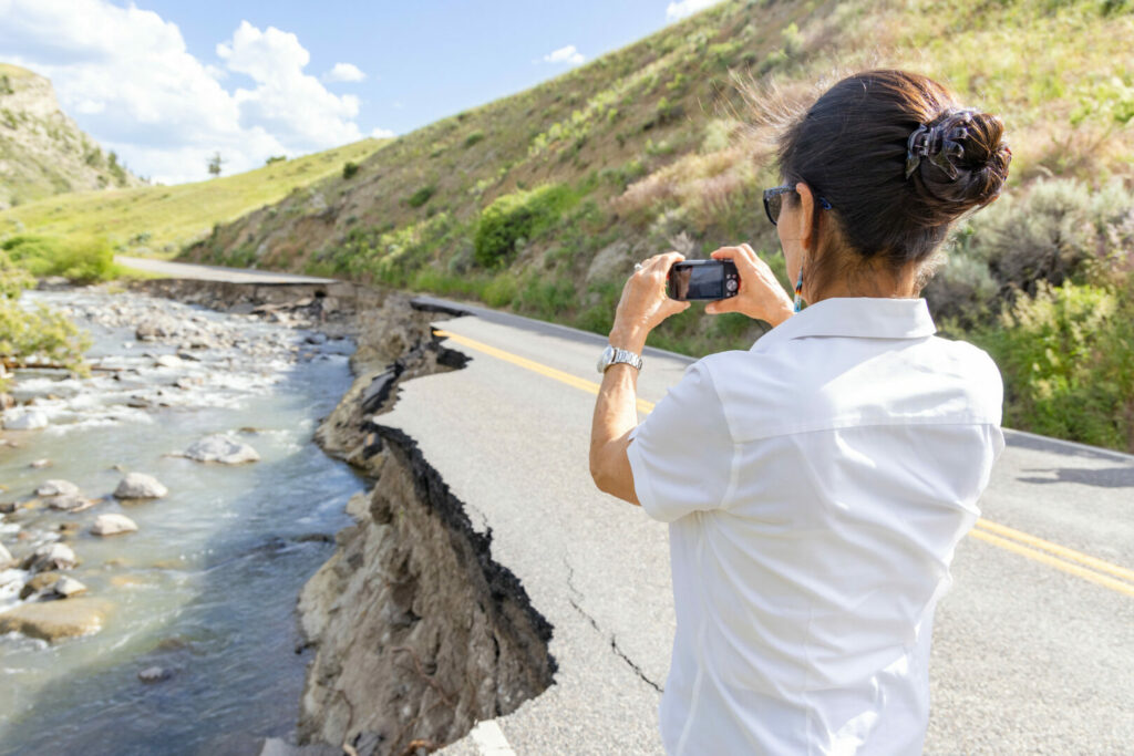 A woman holding a camera up to take a picture of the road that has been completely washed out by flooding in Yellowstone National Park