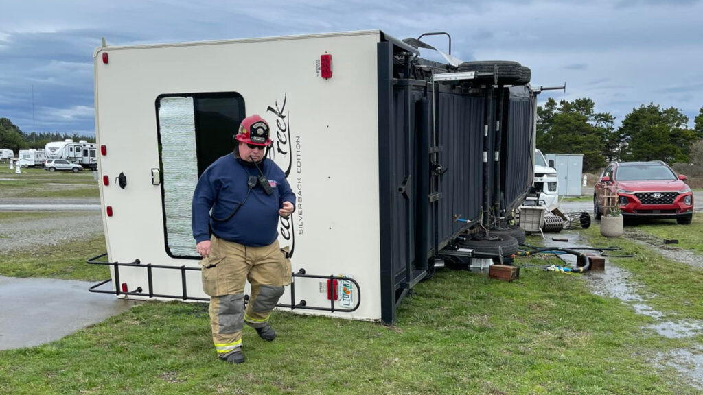 A firefighter walking around an RV that has flipped on its side in the wind 