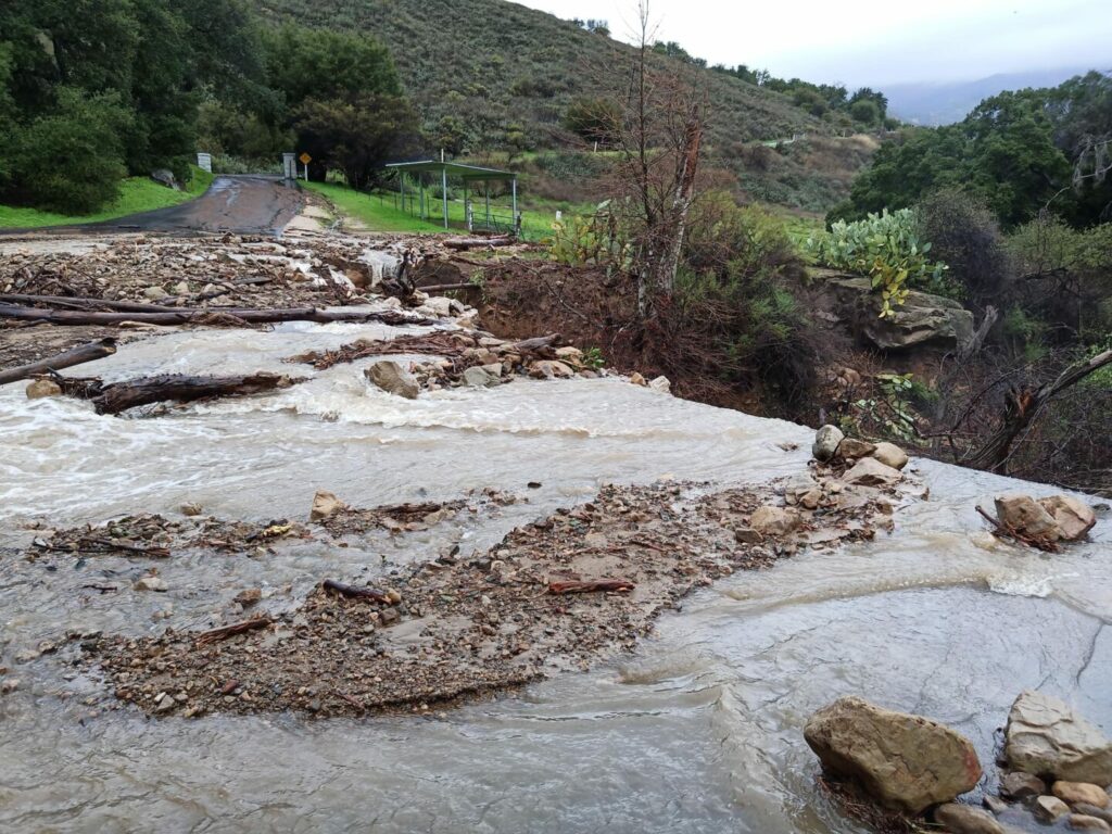 Road leading out of Rancho Oso RV Resort with a portion completely covered in flood water.
