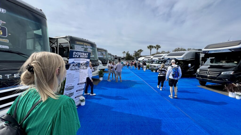 A woman standing in an RV show with a row of RVs and people walking. She's learning how to negotiate rv price. 