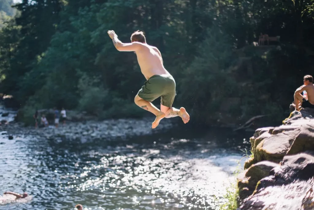 A man jumps off a rock into a river at the base of a waterfall running through the woods in the summertime.