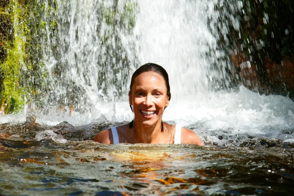 A smiling woman with the sun on her face swims at the base of the falls in a river.
