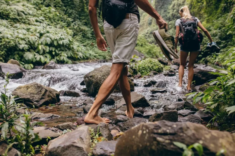 Two hikers take off their shoes to hike through a stream in search of a waterfall.