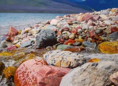 The colorful rocky shoreline of Lake McDonald with tall blue mountains across the lake.