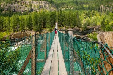 A man stands in the middle of a swinging bridge in Montana that surrounded by lush green forests and beautiful blue water below.