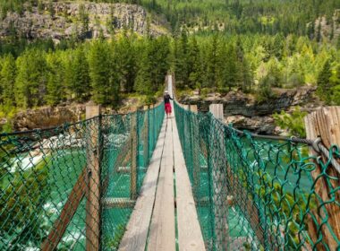 A man stands in the middle of a swinging bridge in Montana that surrounded by lush green forests and beautiful blue water below.