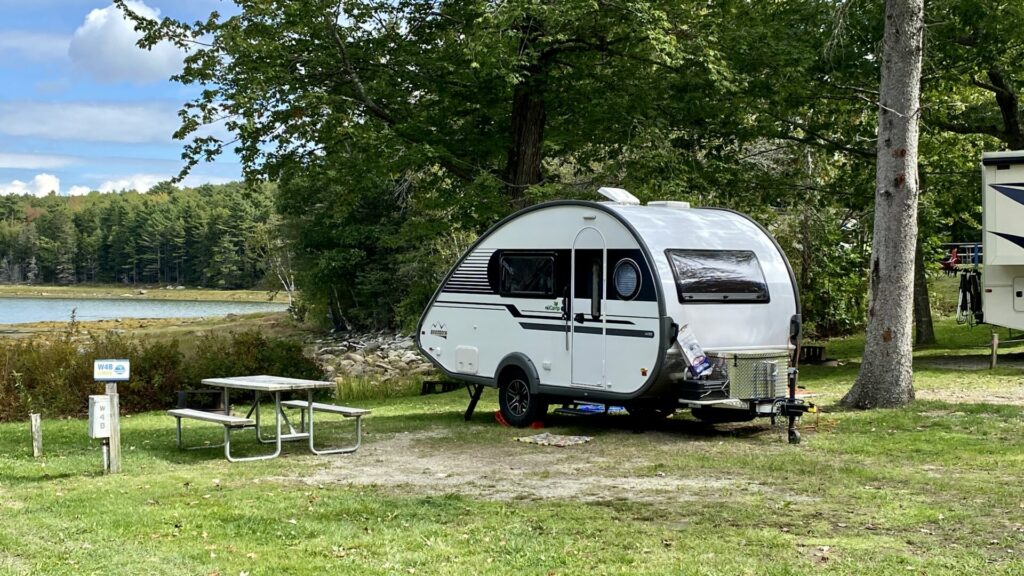 A small travel trailer parked in a campsite with a picnic table and view of a lake. 