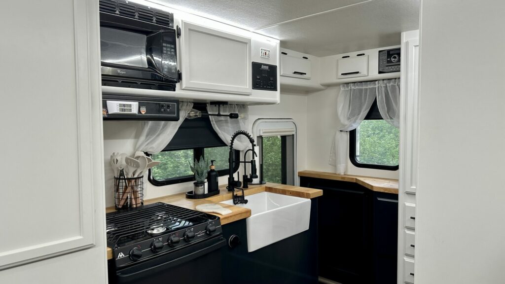 Our RV kitchen after the renovation showing black cabinets and white cupboards, farmhouse style sink, new faucets, and butcher block counter tops. 